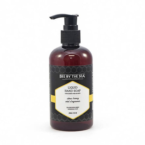 A bottle of Bee By The Sea Liquid Hand Soap in citrus honey and mid-agrumes scent, featuring a dark amber bottle, black pump, and a black label with a yellow and white geometric design.